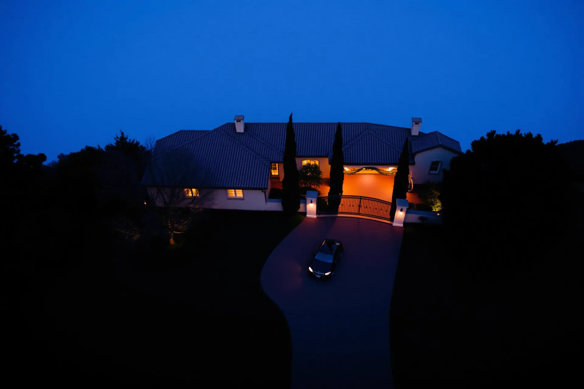 Aerial view at blue hour of a Mediterranean estate driveway and gated entrance, warm gate lights glowing. (AI-generated image)