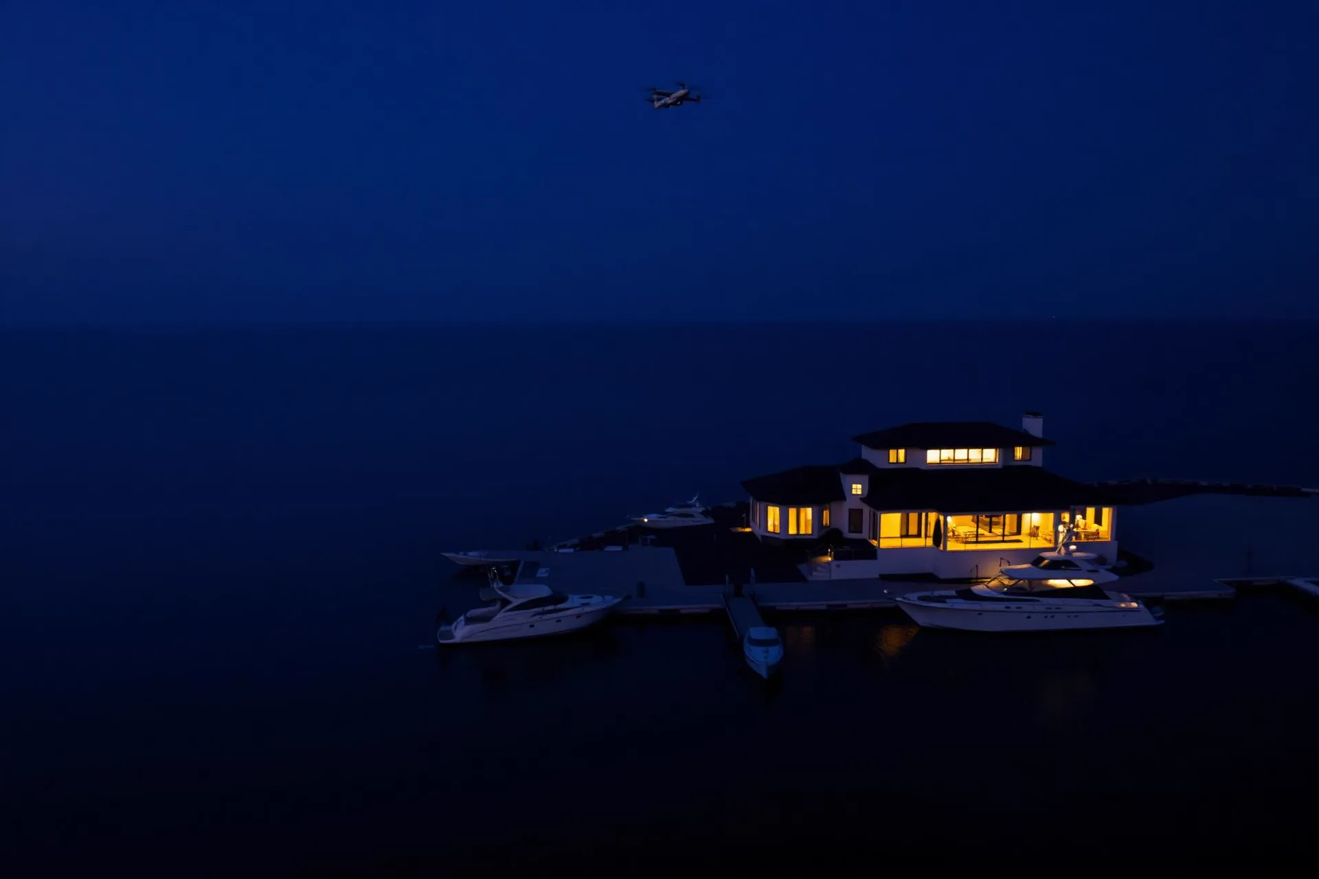 Aerial view at night of a marina-side residence with moored yachts, a faint drone silhouette overhead. (AI-generated image)