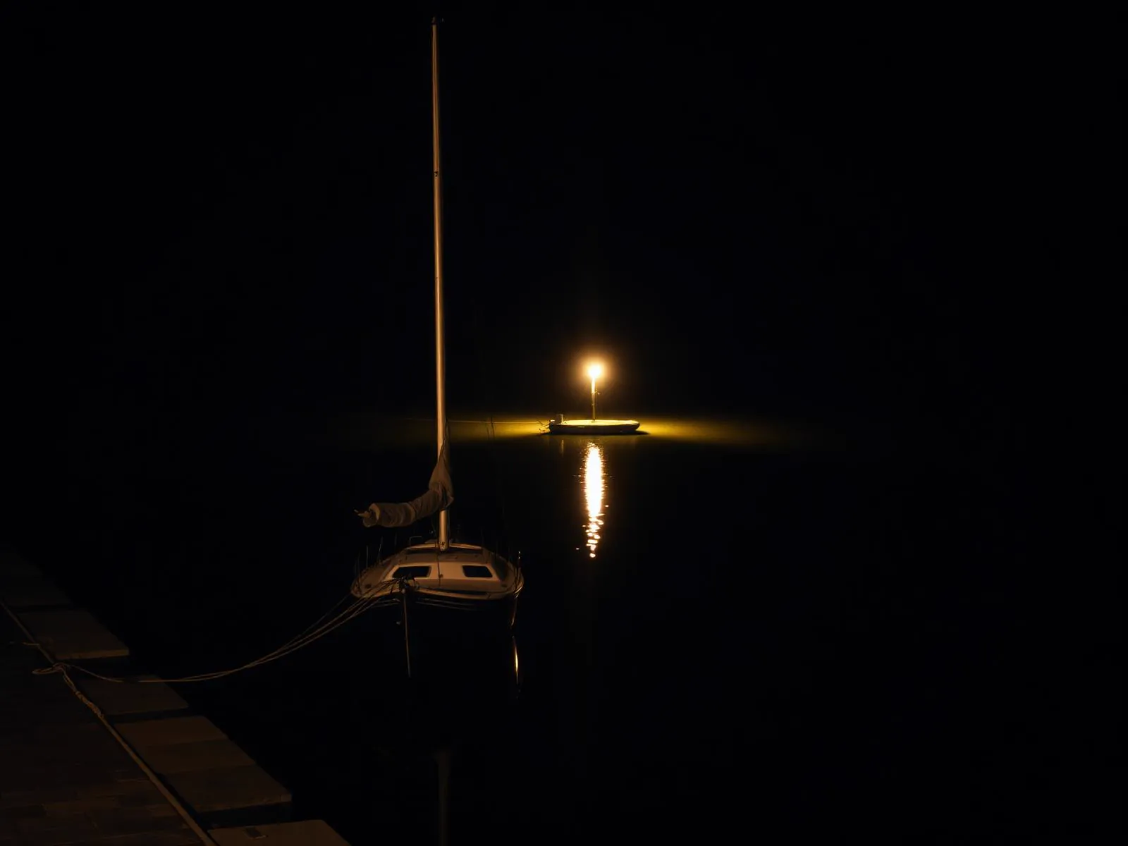 A lone moored sailboat by a stone quay at night, single warm dock lamp reflected on the water. (AI-generated image)